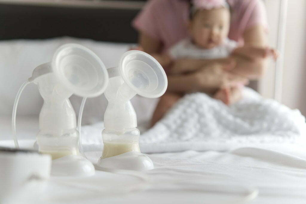 Breast milk bottles and pump machine on the bed with mother hugging baby in background.